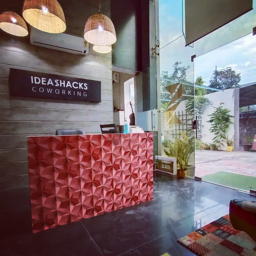 Reception area of IDEASHACKS Coworking with a red geometric-textured desk, wicker pendant lights, and a glass door opening to a greenery-filled outdoor space